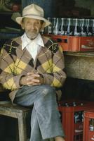 An old man in a hat sitting in front of a case of empty coke bottles