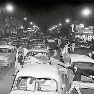 A long line of students holding hands weaving around multiple lanes of cars on a busy street at night. Photo taken on the 200 block of 2nd Ave S in Saskatoon in the 1950's.